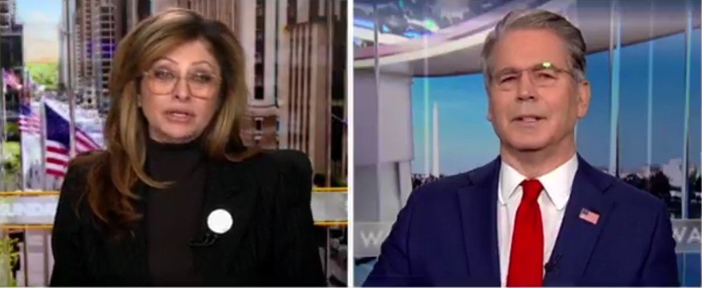 Two news anchors discussing current events, with an urban background featuring flags and a modern studio setting.