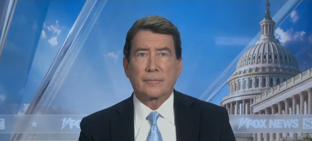 News anchor presenting in front of the U.S. Capitol, featuring a professional appearance with a blue tie and a serious expression.