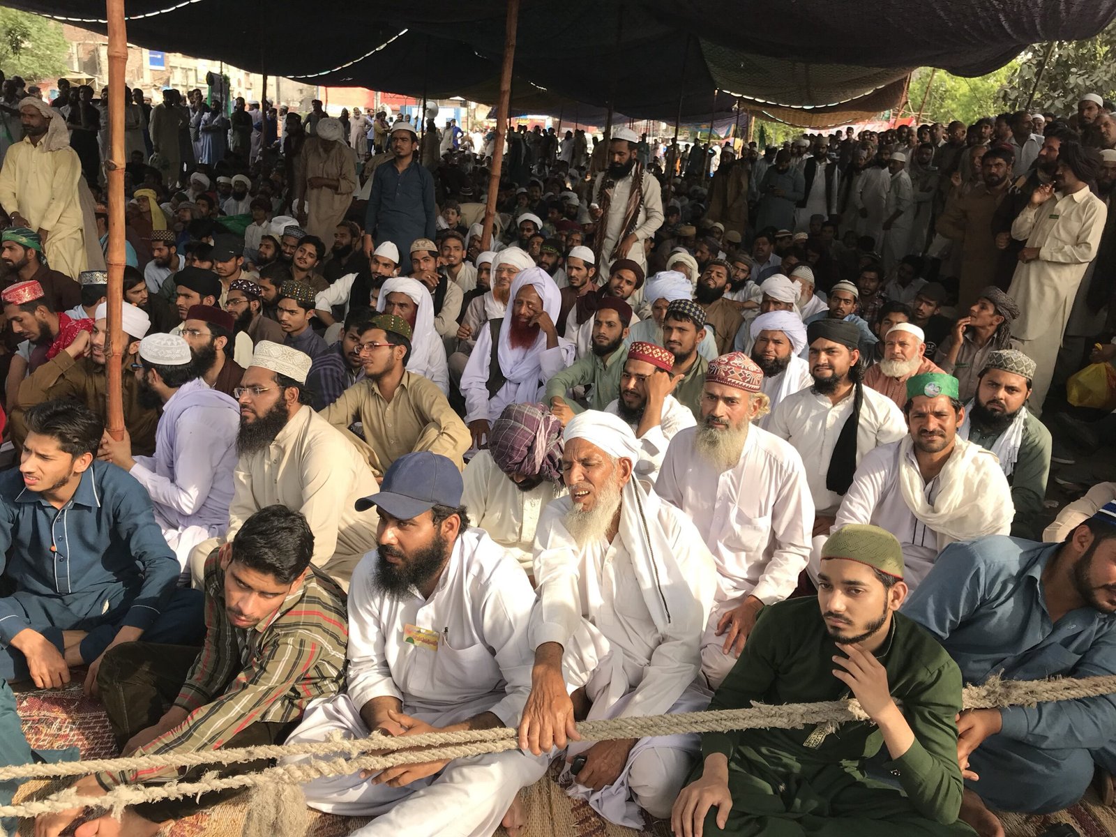 A diverse group of men sitting together in a large gathering, wearing traditional attire and head coverings, engaged in a communal event under a shaded tent.
