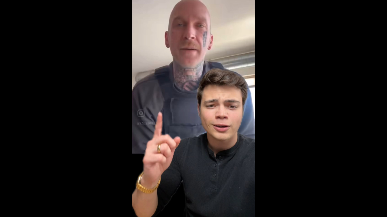 Two men engaged in conversation, one wearing a tactical vest with tattoos, while the other gestures with his hand, indicating a point of interest.