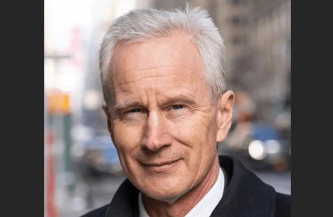 Portrait of a mature man with gray hair and a confident smile, set against an urban backdrop with blurred traffic in the background.