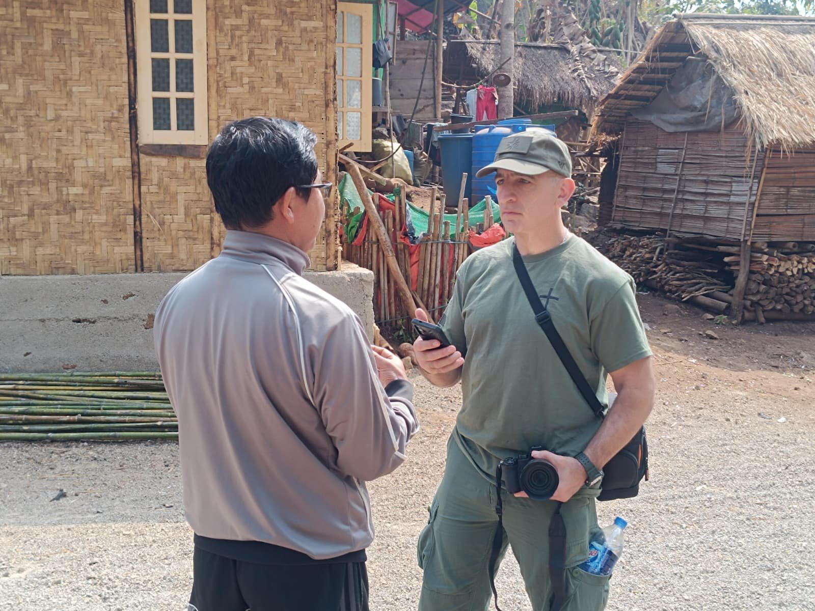 Two men engage in conversation outside a rural village, with one holding a smartphone and the other wearing sunglasses, amid traditional thatched houses and natural scenery.