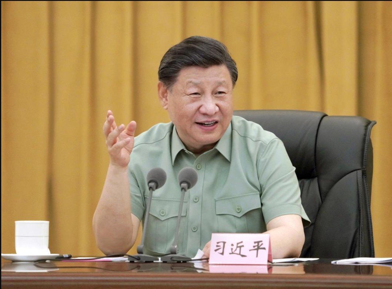Chinese President Xi Jinping speaking during a meeting, gesturing with his hand, seated at a table with a cup and documents, against a backdrop of golden curtains.