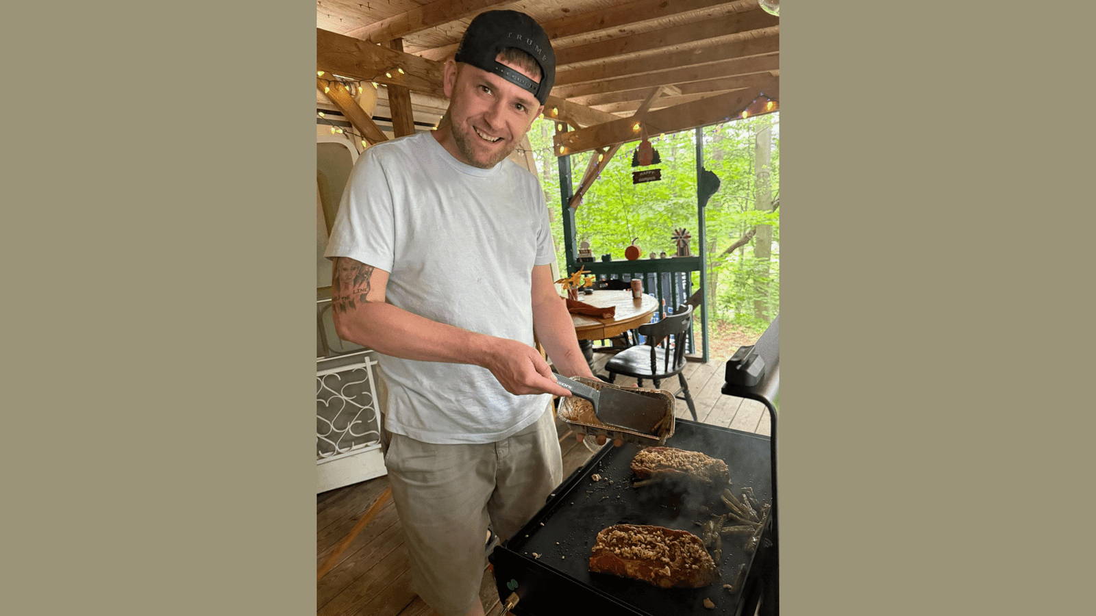 Man grilling seasoned meat and vegetables on a covered porch surrounded by greenery, showcasing outdoor cooking and a relaxed atmosphere.